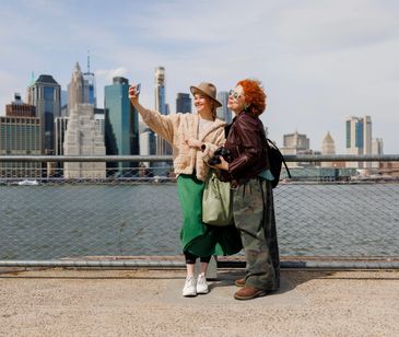 Two friends taking a selfie by the waterfront with city skyline behind them.