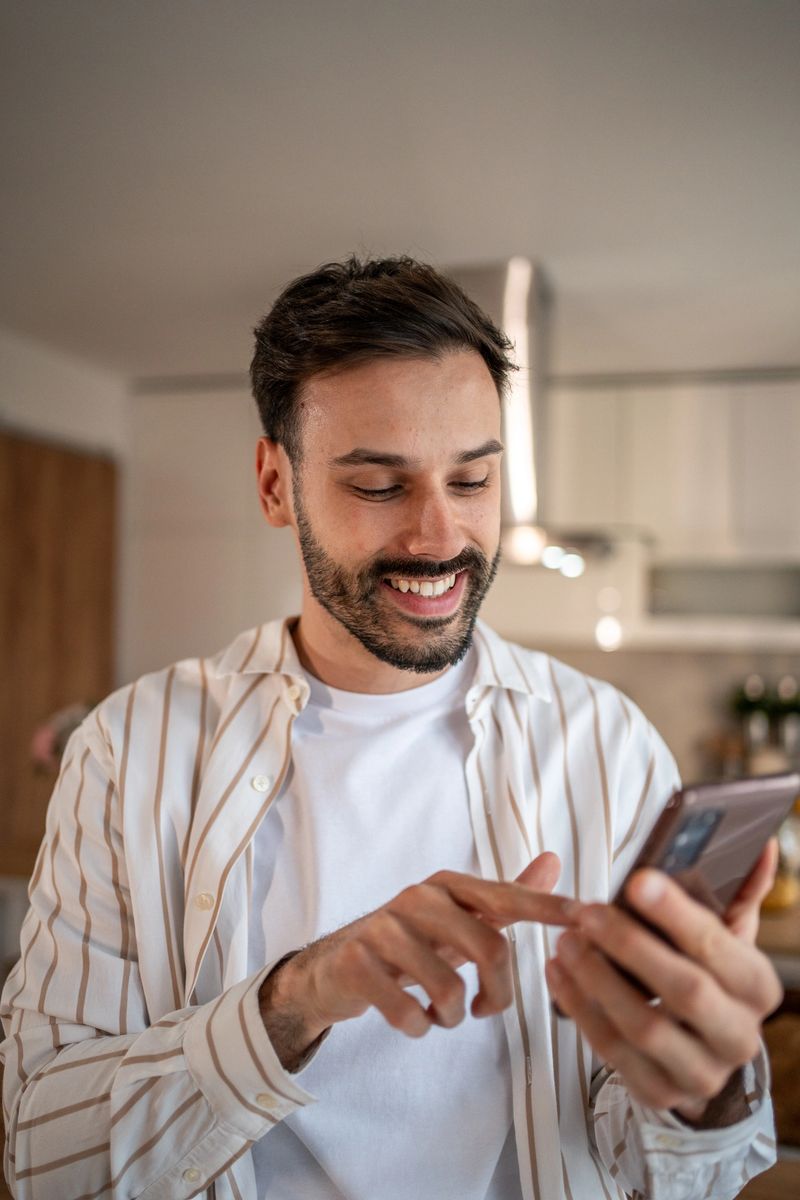 Young man standing in a modern kitchen, smiling while using his smartphone, enjoying a moment of connection and communication in a bright, comfortable home environment