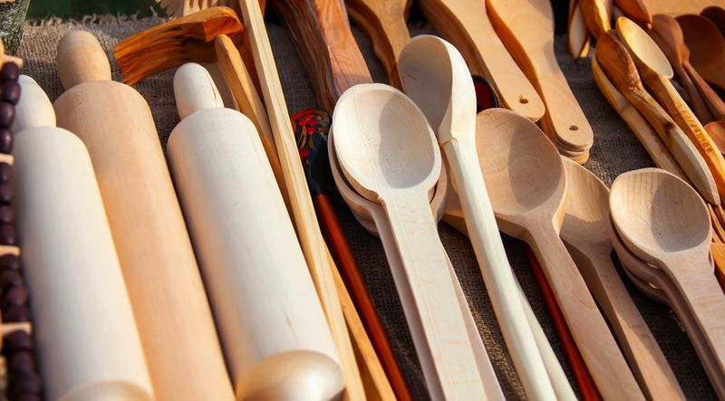 A collection of handcrafted wooden utensils including rolling pins and spoons is arranged on a table at an artisan market, showcasing craftsmanship.