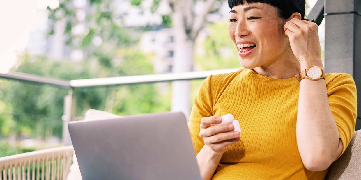 Woman smiling while using wireless earbuds and laptop outdoors.