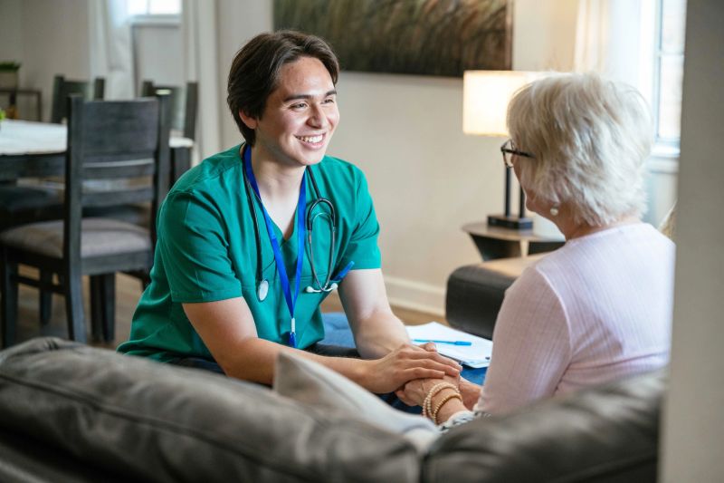 Young male nurse in green scrubs sitting and holding hands with elderly woman in a cozy living room setting