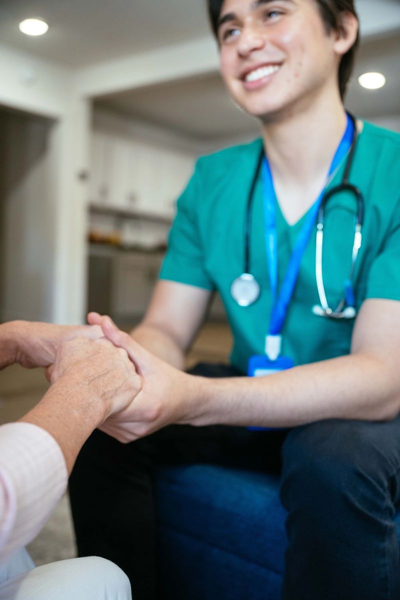 Young male nurse in green scrubs sitting and holding hands with elderly woman in a cozy living room setting