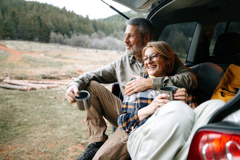Smiling couple of hikers relaxing at the open trunk of their car, sipping hot drinks while surrounded by nature. Peaceful break after an outdoor adventure