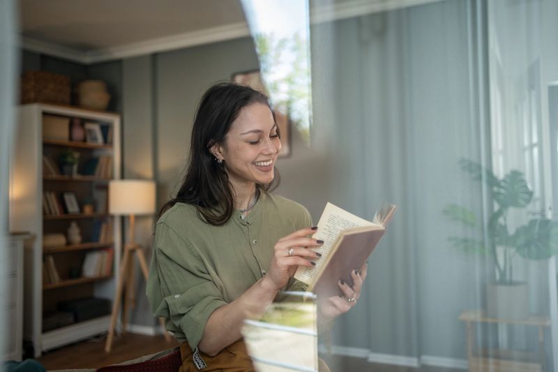 Smiling young woman immersed in a book while sitting comfortably in her living room, savoring a peaceful moment of leisure and relaxation amid cozy surroundings