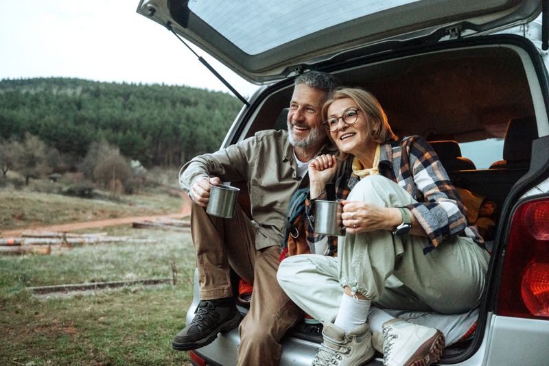 Cheerful mature hikers enjoying hot coffee while sitting on the edge of their car’s open trunk. Relaxing together in a beautiful natural setting.