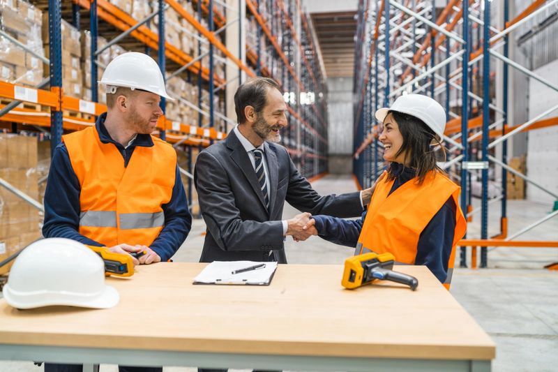 Manager and storage worker shaking hands after finalizing a contract in a bustling warehouse, highlighting teamwork and successful collaboration