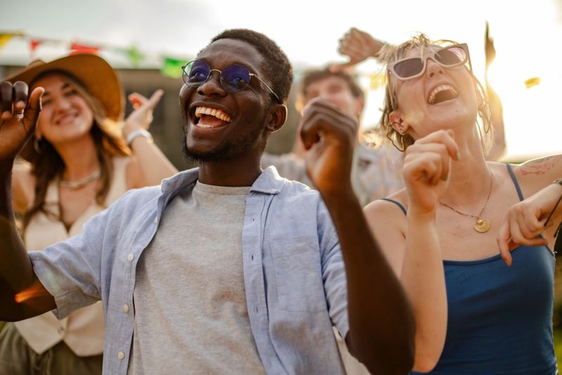 A diverse crowd of young people enjoys music, dancing, and laughter at an outdoor festival. With arms raised, they celebrate friendship and happiness during the sunset.