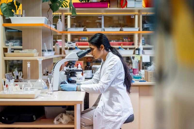 Close-up portrait of young South Asian female scientist in white lab coat performing microscopic analysis in contemporary research facility with organized laboratory equipment and supplies.