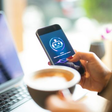 Person using chatbot app on smartphone with coffee and laptop nearby.