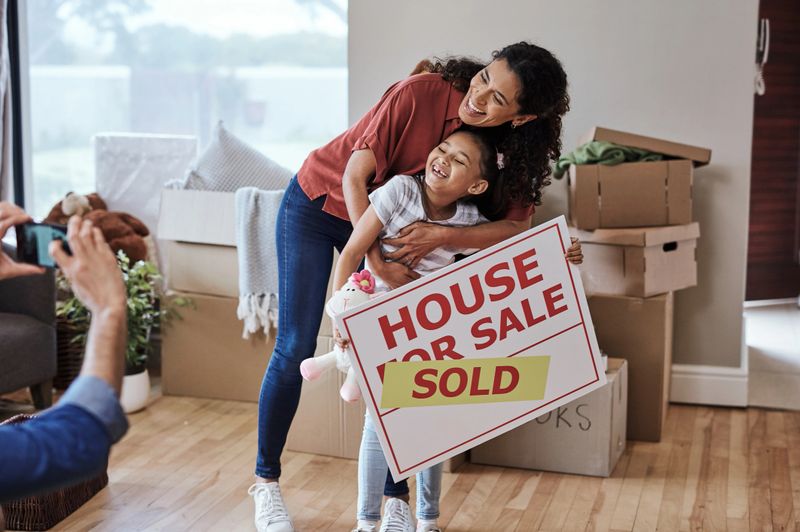A joyful mother and daughter enjoy a special moment in their new home, posing with a 'House For Sale - Sold' sign amidst moving boxes, portraying happiness and fresh beginnings.