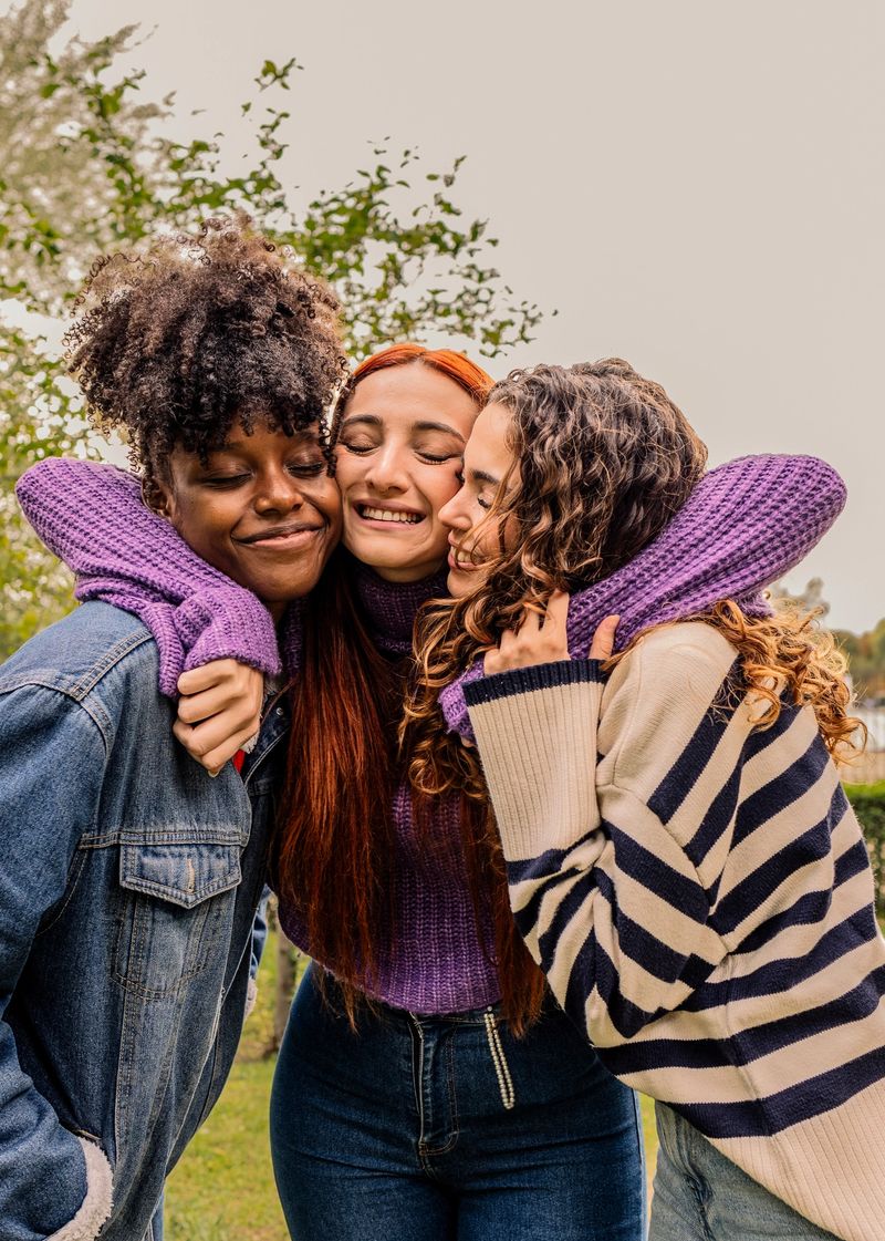 Three cheerful young women, representing different ethnicities, are hugging tightly in a park, expressing their strong bond and celebrating their friendship with joy and affection