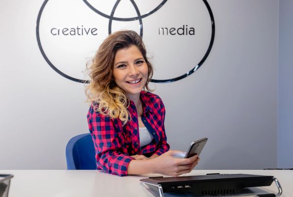Smiling woman holding phone at desk with creative media wall art.