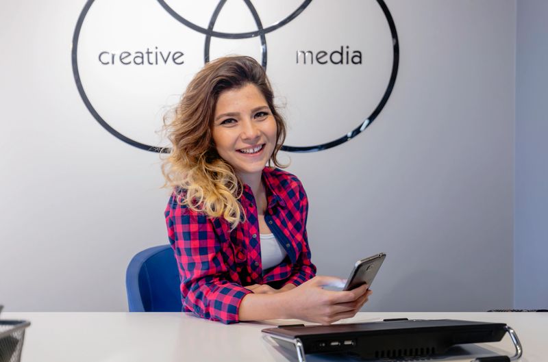 A cheerful young woman with wavy ombre hair, dressed in a red plaid shirt and white top, is seated at a sleek white desk inside a modern creative media office. She holds a smartphone in one hand and smiles warmly toward the camera, suggesting confidence, approachability, and digital fluency. The wall behind her displays overlapping black circles labeled "creative" and "media," hinting at a dynamic agency or content studio setting. The scene evokes a blend of youthful energy, professionalism, and social media engagement in a startup or freelance work environment