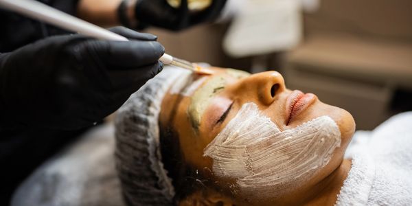 A woman is receiving a facial treatment with a brush applying a mask.