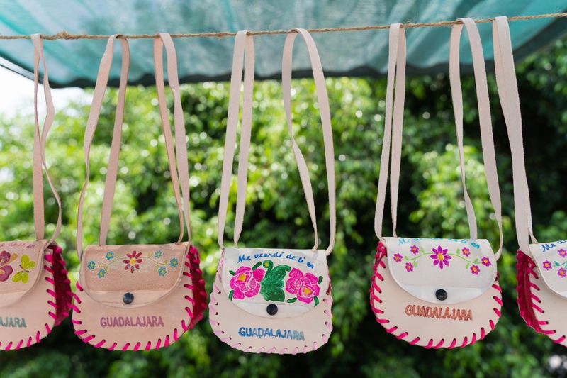 Handcrafted souvenir leather bags with floral embroidery and "Guadalajara" text hanging on display at a local market in Jalisco, Mexico. Language translation: I thought of you in Guadalajara