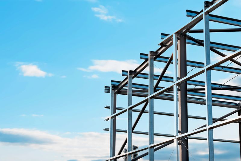 A steel framework is being erected in an urban location under a bright blue sky with scattered clouds, illustrating progress in modern building construction.