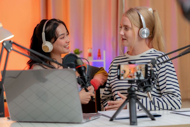 Confident women hosting a podcast while live streaming from a studio with microphones, headphones, and sound equipment. Smiling warmly as they connect with a remote audience, showcasing teamwork, creativity, journalism, and modern digital media culture.