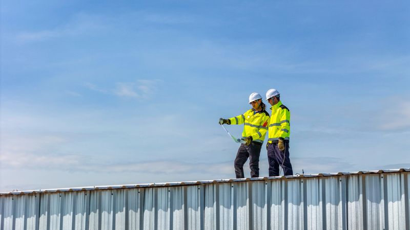 Two engineers in safety gear standing on a rooftop reviewing blueprints for a solar panel installation project, showcasing teamwork, renewable energy planning, and sustainable construction practices.