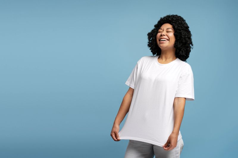 Happy young African American woman with natural curls laughing, wearing white t-shirt and standing against blue studio. background Lifestyle, diversity and emotions concept