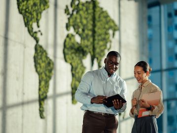 Two professionals discussing information on a tablet in an office with a green world map wall.