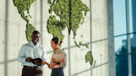 Two professionals discussing work with a tablet in front of a world map wall.