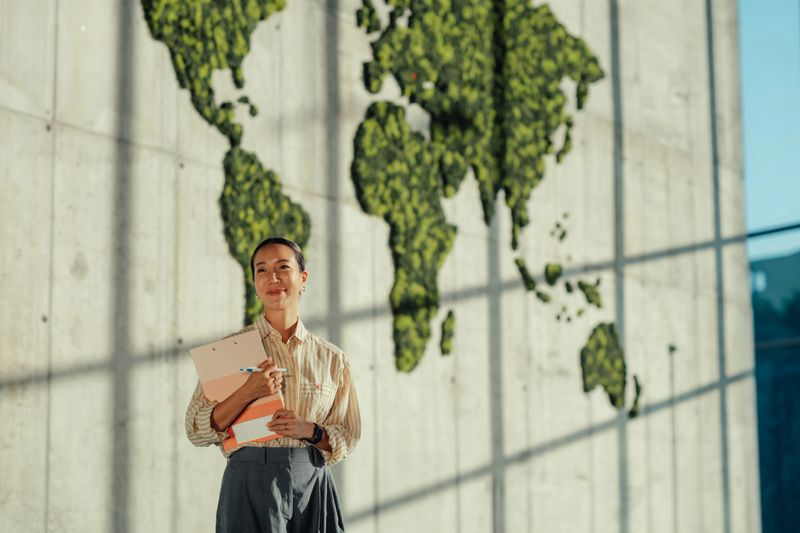 Portrait of a businesswoman holding a clipboard in a modern office with a green world map in the background, symbolizing global business and environmental awareness