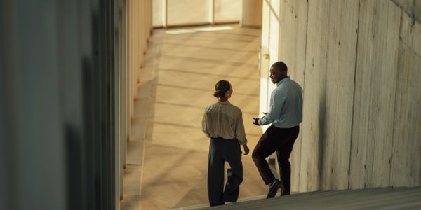 Two professionals walking and talking in a modern, sunlit hallway.