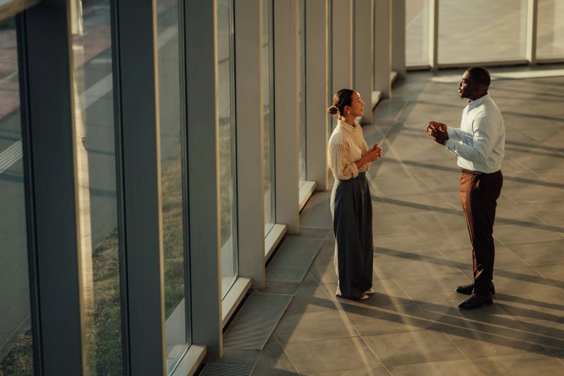 Two businesspeople are having a discussion near a large window in a modern office building, illuminated by warm sunlight
