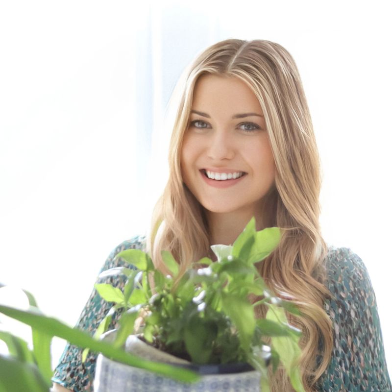Woman smiling with long blonde hair, wearing a green dress and holding a potted houseplant, standing in a bright home interior filled with sunlight