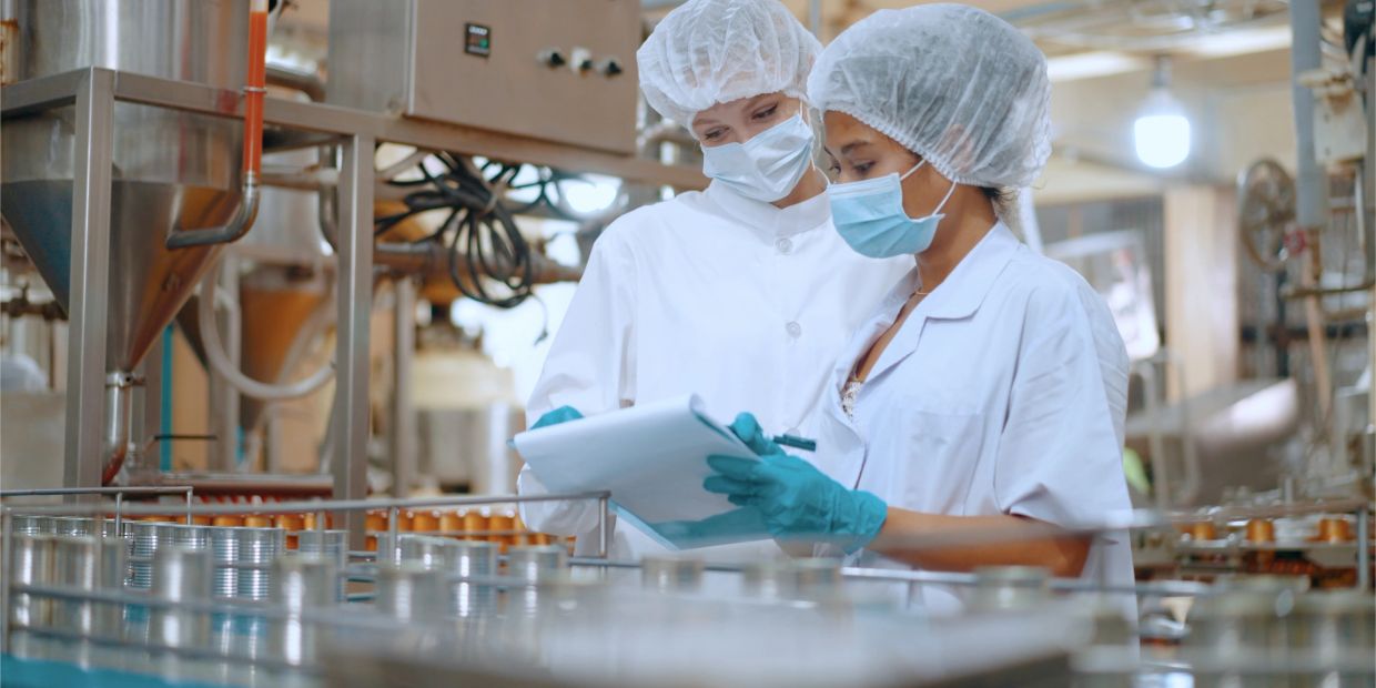 Two women in protective gear inspecting cans in a factory.