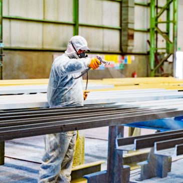 Worker spraying paint on metal beams in protective gear.