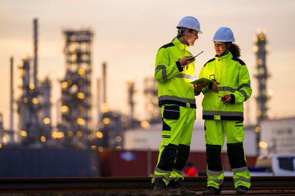 Two workers in high-visibility gear discuss plans at an industrial site during sunset.