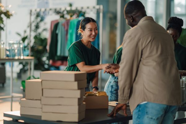 A woman smiles and shakes hands with a man at a table with stacked boxes.
