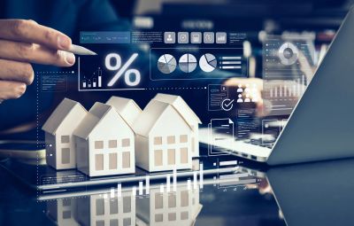 Hand pointing at digital financial charts above model houses and laptop.