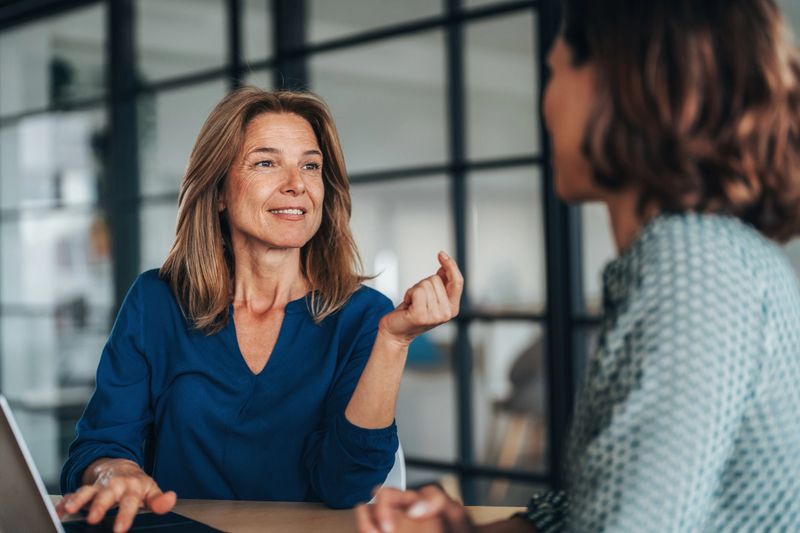 Two women having a focused and friendly conversation at a desk. They wearing formal businesswear, smiling and gesturing with hands, explaining something in a discussion. One woman talking and The other woman, listens attentively while using a laptop. Mentorship, collaboration, or client consultation.
