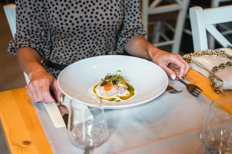 Close-up photograph of an elegantly plated gourmet dish served on pristine white porcelain, accompanied by crystal stemware on a wooden dining table.