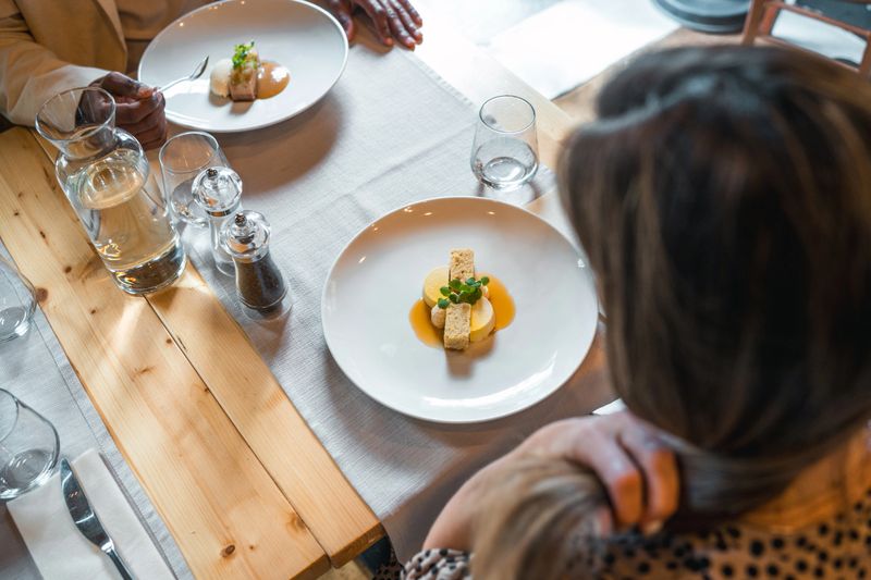 Overhead view of an upscale restaurant interior featuring minimalist white plates with precisely plated gourmet dish, set on rustic wooden table with crystal glassware.