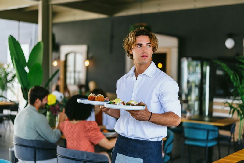 A waiter carries a plate of appetizing dishes while a group of friends enjoys a meal in a lively restaurant setting. The image captures the essence of dining, companionship, and relaxation in a vibrant atmosphere.