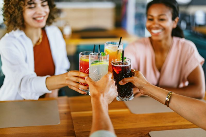 A group of friends joyfully toast with colorful non-alcoholic drinks while enjoying a casual gathering at a restaurant. Smiles and laughter emphasize the sense of togetherness and fun among the diverse group.
