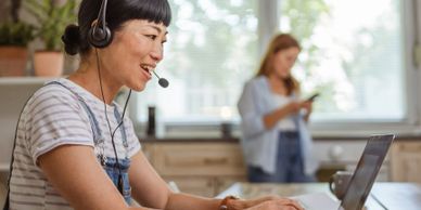 Woman wearing headset working on laptop in a bright home office.