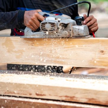 Close-up of hands using an electric planer on wood, with wood shavings flying.
