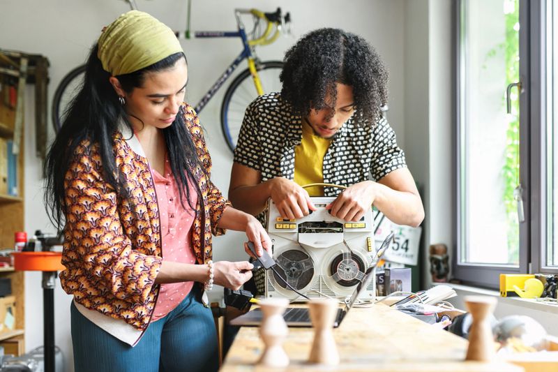 young woman and man connecting retro reel to reel tape recorder with laptop at wooden desk in workshop