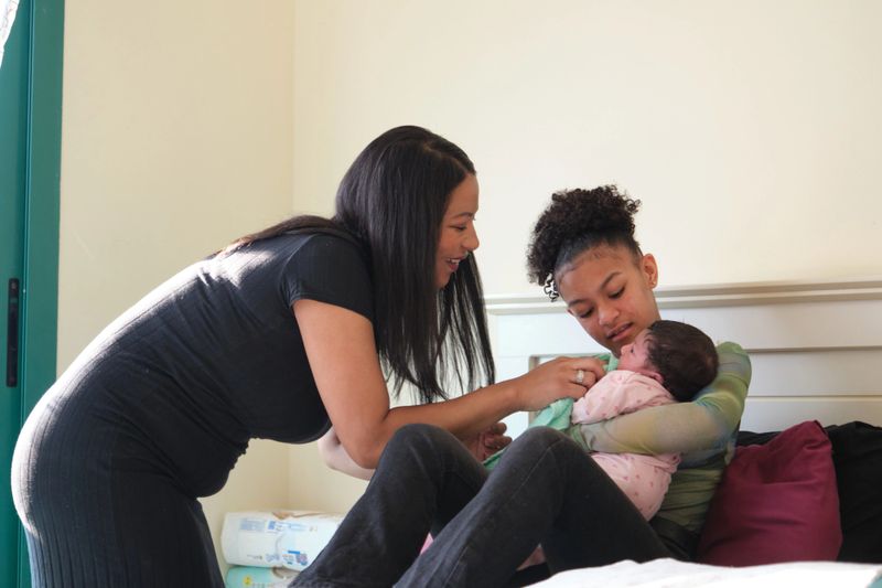Smiling woman playing with her newborn daughter at home. Family is spending leisure time with their infant. They are symbolizing togetherness.