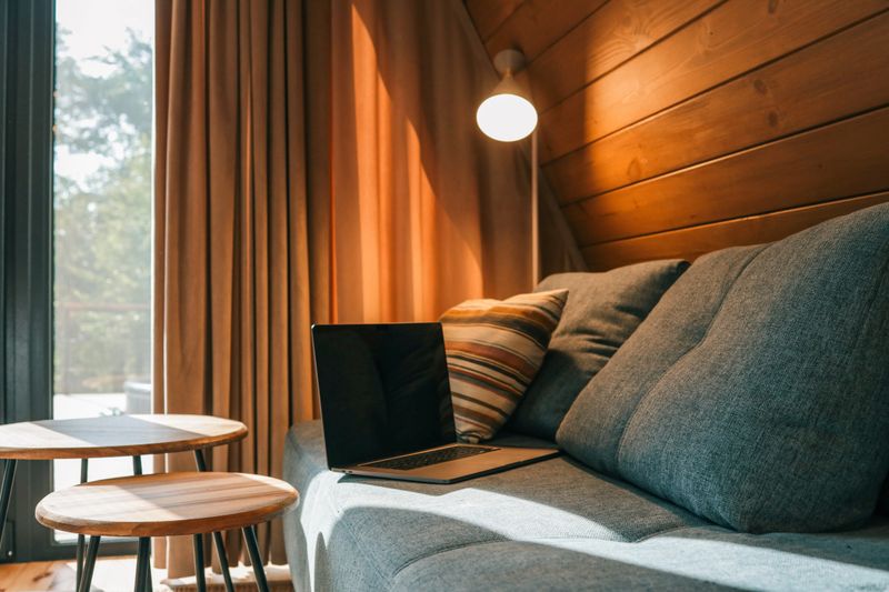 Laptop on a blue couch in a cozy wooden living room with warm natural light, striped pillow, round coffee tables, and a floor lamp near the curtain.
