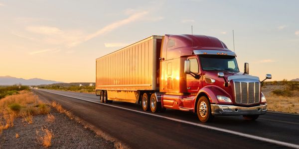 Red semi-truck driving on a desert highway at sunset.