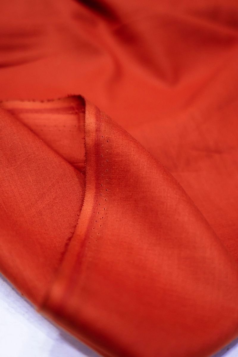 Close-up view of a richly textured, burnt orange fabric. The folds and drapes create a sense of luxury and elegance. The subtle lighting enhances the depth of the color and the texture of the material. The setting appears to be a studio or workshop, with a hint of a light-colored surface visible at the bottom.