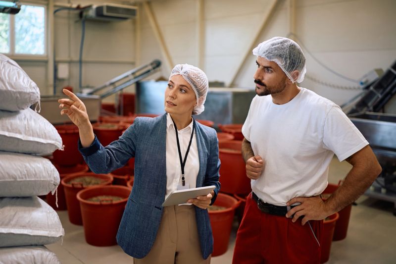 Quality control manager talking to a worker while inspecting processed goods in food factory.