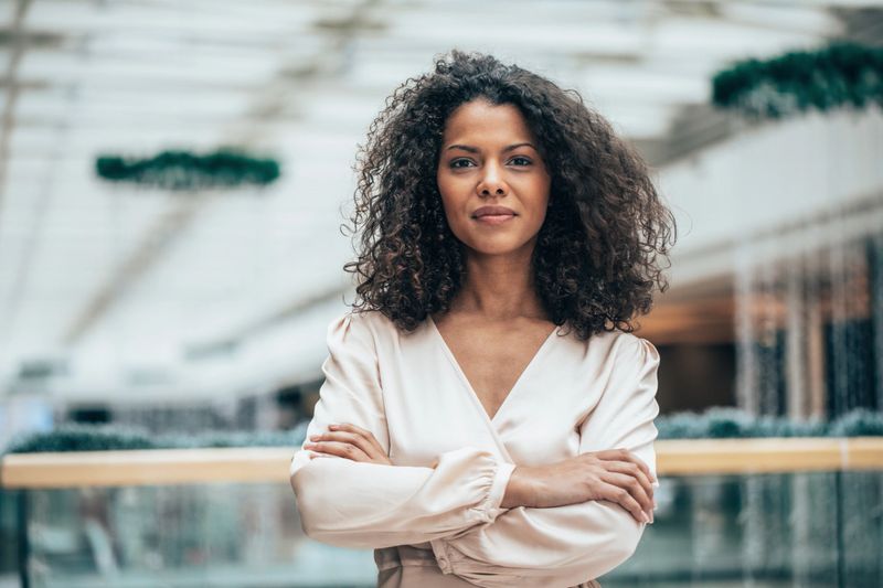 Beautiful afro-american businesswoman. A professional woman standing with crossed arms in a modern office environment. She is looking at camera and smiling. She exudes confidence and leadership, wearing a stylish blouse