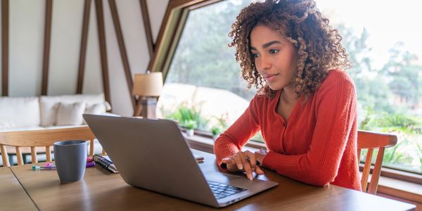 Woman in red sweater working on laptop at wooden table near large window.