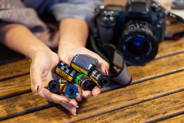 Hands holding several rolls of camera film with a DSLR camera in the background.